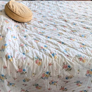 Puede incluir: Un edred&oacute;n acolchado con un estampado floral en colores pastel rosa, azul y amarillo. Una almohada ovalada de color beige descansa sobre el edred&oacute;n. El edred&oacute;n tiene un patr&oacute;n de costura en forma de diamante.