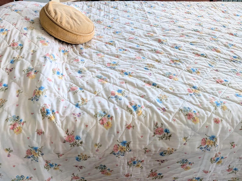 Puede incluir: Un edred&oacute;n acolchado con un estampado floral en colores pastel rosa, azul y amarillo. Una almohada ovalada de color beige descansa sobre el edred&oacute;n. El edred&oacute;n tiene un patr&oacute;n de costura en forma de diamante.