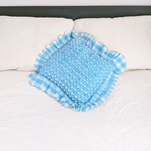 Op de afbeelding: Een blauw en wit geruit kussen met een gerimpelde rand ligt op een wit bed. Het kussen is gevuld met een blauw, gestikt materiaal.