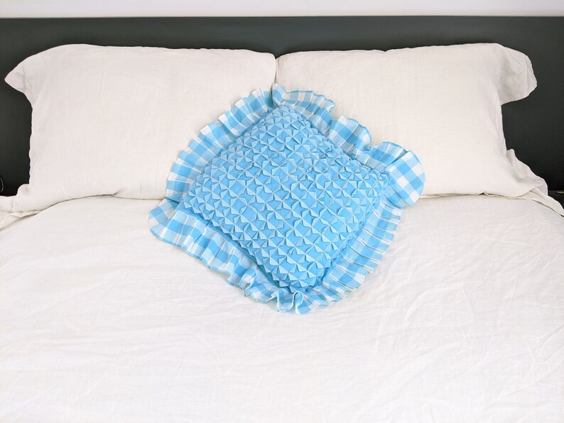 Op de afbeelding: Een blauw en wit geruit kussen met een gerimpelde rand ligt op een wit bed. Het kussen is gevuld met een blauw, gestikt materiaal.