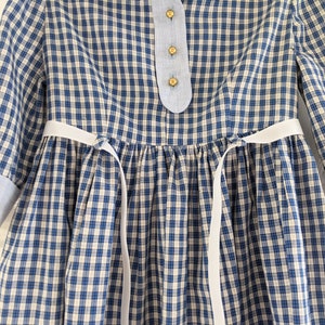 Op de afbeelding: Een blauw-wit geblokt jurkje met witte lintjes aan de taille. Het jurkje heeft een blauwe kraag en knoopsluiting.