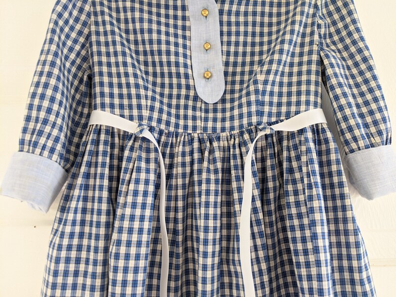Op de afbeelding: Een blauw-wit geblokt jurkje met witte lintjes aan de taille. Het jurkje heeft een blauwe kraag en knoopsluiting.