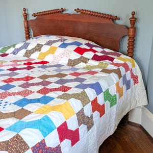May include: A patchwork quilt featuring a repeating bow-tie pattern made of various colorful cotton fabrics in red, blue, yellow, green, and brown, interspersed with white fabric blocks, draped over a wooden headboard with turned spindle posts.