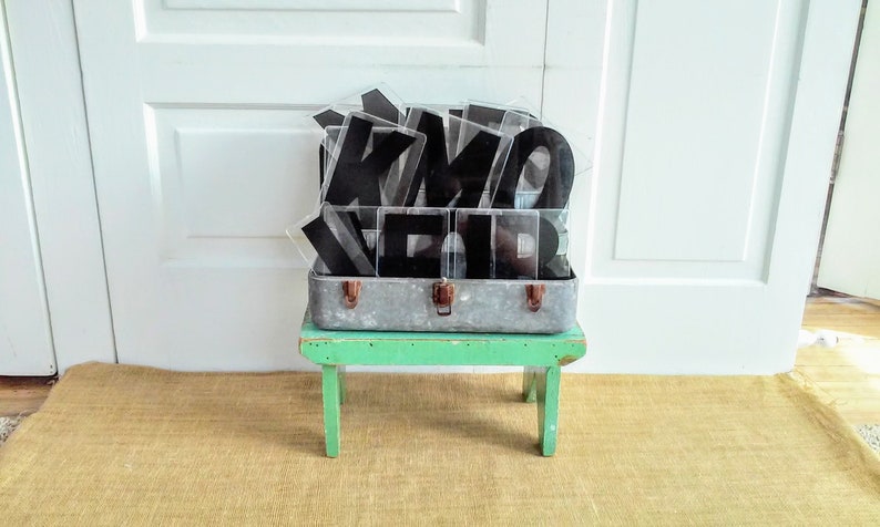 May include: A vintage metal suitcase filled with black plastic letters, sitting on a green wooden stool. The suitcase and letters are on a brown woven rug.