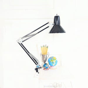 May include: A black and silver articulated desk lamp with a conical shade is clamped to a white surface. A small globe, pencils, and a trophy-like container are on the surface below the lamp.