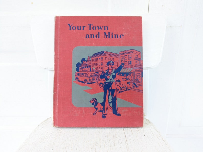 May include: A red hardcover book with the title "Your Town and Mine" in blue lettering. The cover features an illustration of a city street scene with a traffic officer directing traffic, cars, and a bus.