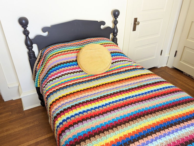 Op de afbeelding: Een zwart houten bedframe met een kleurrijk gestreept gehaakt deken en een rond bruin kussen.