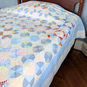 Puede incluir: Un edred&oacute;n hecho a mano con un patr&oacute;n de molinillo en colores pastel de azul, rosa, naranja y verde. El edred&oacute;n est&aacute; sobre una cama con cabecero de madera. El edred&oacute;n tiene un borde azul claro y un fondo color crema.