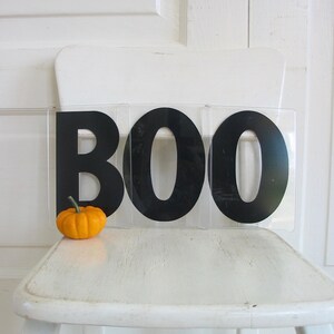 May include: A white chair with a black BOO sign on it. A small orange pumpkin sits on the chair in front of the sign.