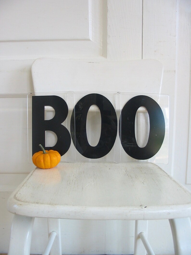 May include: A white chair with a black BOO sign on it. A small orange pumpkin sits on the chair in front of the sign.