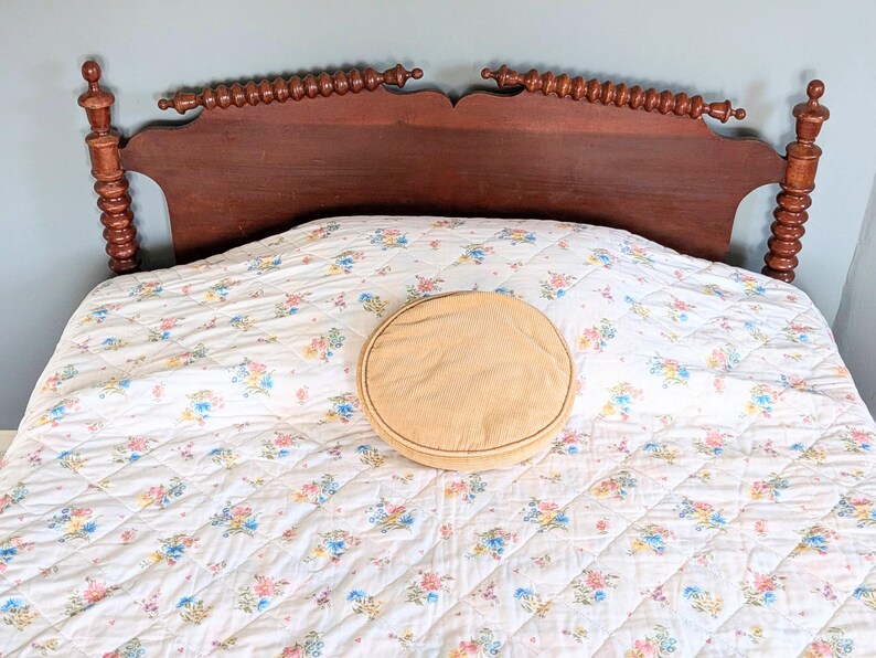 Puede incluir: Un marco de cama de madera vintage con acabado marr&oacute;n oscuro. El cabecero presenta husillos decorativos y una parte superior curvada. La cama est&aacute; hecha con un edred&oacute;n con estampado floral y una almohada redonda y beige.