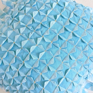 Op de afbeelding: Een lichtblauw kussen met een gestikt ontwerp met een herhalend patroon van kleine, gevouwen vierkanten. De vierkanten zijn gerangschikt in een rasterpatroon en zijn geaccentueerd met kleine, goudkleurige stippen.