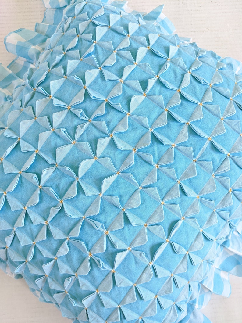 Op de afbeelding: Een lichtblauw kussen met een gestikt ontwerp met een herhalend patroon van kleine, gevouwen vierkanten. De vierkanten zijn gerangschikt in een rasterpatroon en zijn geaccentueerd met kleine, goudkleurige stippen.