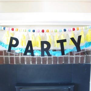 May include: A black and white party banner with the word "PARTY" in large, bold letters. The banner is decorated with yellow and blue tissue paper tassels and colorful beads.