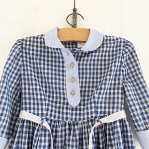 Op de afbeelding: Een blauw-wit geblokt jurkje met een witte strik om de taille. Het jurkje heeft een Peter Pan-kraag en lange mouwen met blauwe manchetten.
