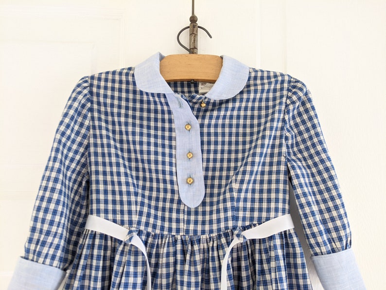 Op de afbeelding: Een blauw-wit geblokt jurkje met een witte strik om de taille. Het jurkje heeft een Peter Pan-kraag en lange mouwen met blauwe manchetten.