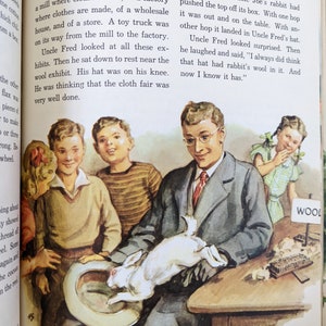 May include: A group of children are looking at a white rabbit sitting on a hat. The rabbit is on a table with a sign that says "Wool" and a comb with wool on it. The children are wearing sweaters and one is wearing a hat.