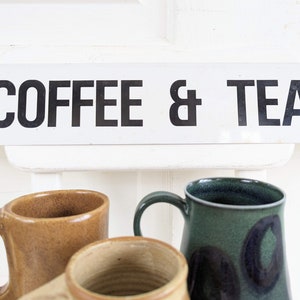 Puede incluir: Un letrero blanco con texto negro que dice "COFFEE & TEA". Tres tazas est&aacute;n en primer plano, una es marr&oacute;n, una es verde y una es beige.