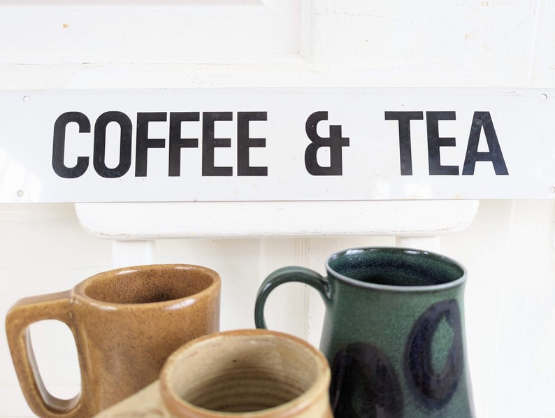 Puede incluir: Un letrero blanco con texto negro que dice "COFFEE & TEA". Tres tazas est&aacute;n en primer plano, una es marr&oacute;n, una es verde y una es beige.