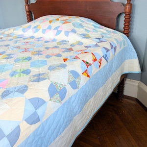 Puede incluir: Un edred&oacute;n vintage con un dise&ntilde;o de patchwork colorido, con formas geom&eacute;tricas azules, rosas y naranjas sobre un fondo crema. El edred&oacute;n tiene un borde azul claro y est&aacute; colocado sobre una estructura de cama de madera.
