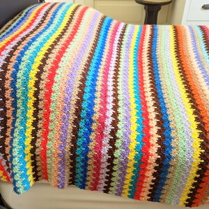 Op de afbeelding: Een kleurrijk gestreept gehaakt deken met verschillende kleuren, waaronder rood, oranje, geel, groen, blauw, paars en bruin.