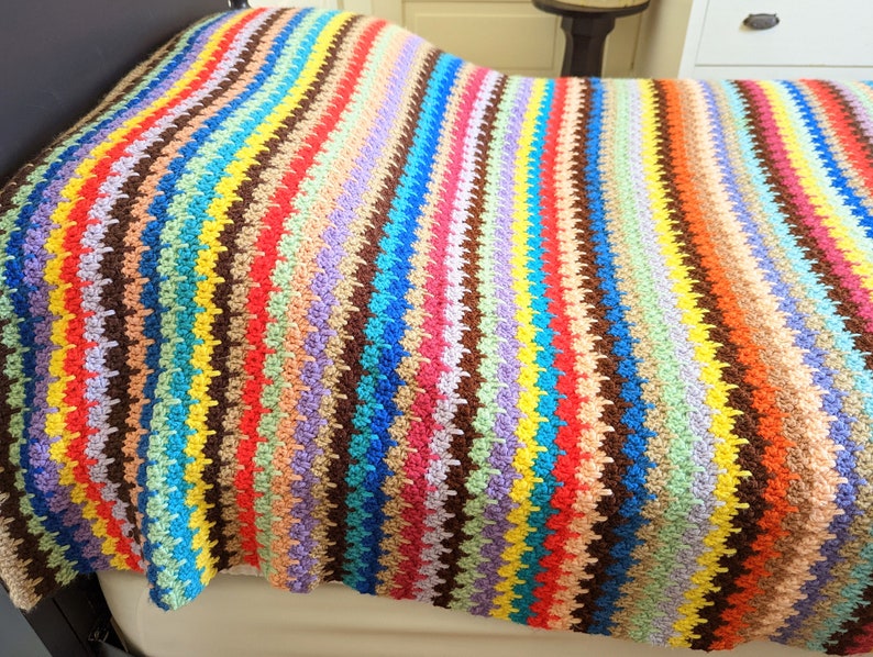 Op de afbeelding: Een kleurrijk gestreept gehaakt deken met verschillende kleuren, waaronder rood, oranje, geel, groen, blauw, paars en bruin.