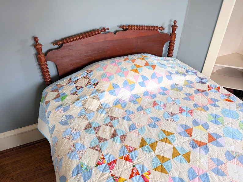 Puede incluir: Una colcha vintage con un colorido patr&oacute;n geom&eacute;trico cubre una cama. La colcha presenta una mezcla de formas azules, rosas, amarillas y naranjas sobre un fondo crema. El cabecero es de madera oscura.