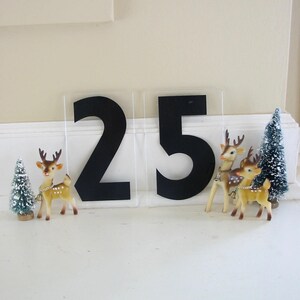 May include: Two black numbers, 25, on a white background with two small, brown, plastic reindeer figurines and a small, green, artificial Christmas tree.
