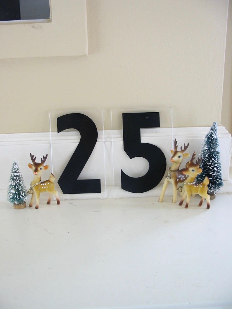 May include: Two black numbers, 25, on a white background with two small, brown, plastic reindeer figurines and a small, green, artificial Christmas tree.