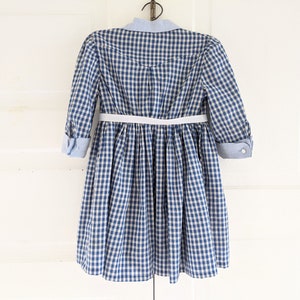 Op de afbeelding: Een blauw-wit gebloemde jurk met een witte riem en een blauwe knoopsluiting.