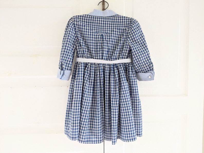 Op de afbeelding: Een blauw-wit gebloemde jurk met een witte riem en een blauwe knoopsluiting.