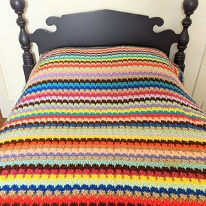 Op de afbeelding: Een kleurrijk gestreept gehaakt deken met een verscheidenheid aan kleuren, waaronder rood, oranje, geel, groen, blauw, paars en bruin. Het deken ligt op een bed met een donker houten hoofdeinde.