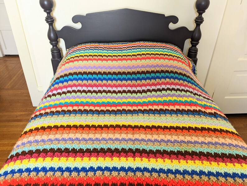 Op de afbeelding: Een kleurrijk gestreept gehaakt deken met een verscheidenheid aan kleuren, waaronder rood, oranje, geel, groen, blauw, paars en bruin. Het deken ligt op een bed met een donker houten hoofdeinde.