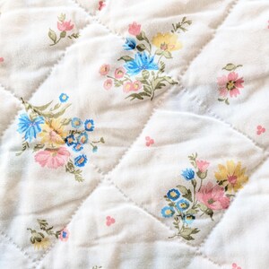 Puede incluir: Primer plano de una tela acolchada con un estampado floral. La tela es blanquecina con un patr&oacute;n de diamantes creado por costuras. Racimos de flores rosas, azules y amarillas est&aacute;n dispersos por la superficie, creando una est&eacute;tica vintage.