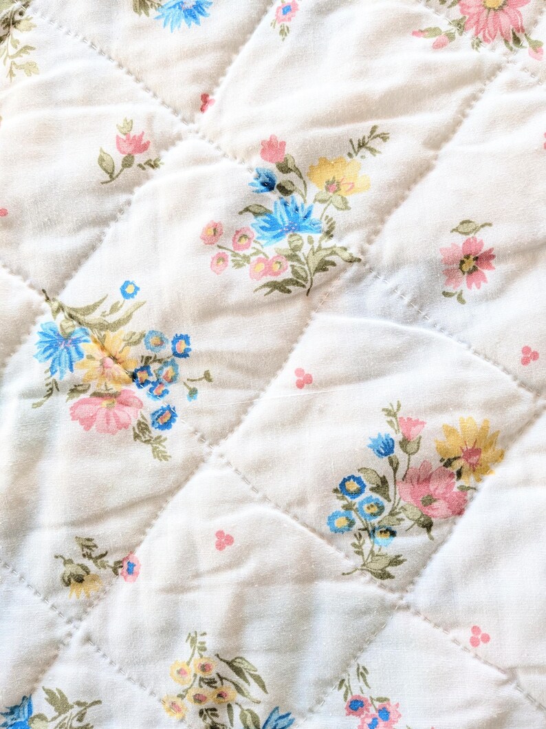 Puede incluir: Primer plano de una tela acolchada con un estampado floral. La tela es blanquecina con un patr&oacute;n de diamantes creado por costuras. Racimos de flores rosas, azules y amarillas est&aacute;n dispersos por la superficie, creando una est&eacute;tica vintage.