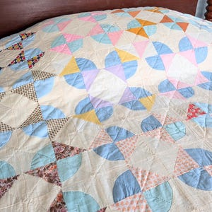 Puede incluir: Un edred&oacute;n hecho a mano con un fondo crema y un patr&oacute;n repetido de formas pastel. El edred&oacute;n presenta arcos y tri&aacute;ngulos azules, rosas, amarillos y naranjas, creando un dise&ntilde;o de patchwork. El edred&oacute;n est&aacute; sobre un marco de cama de madera.