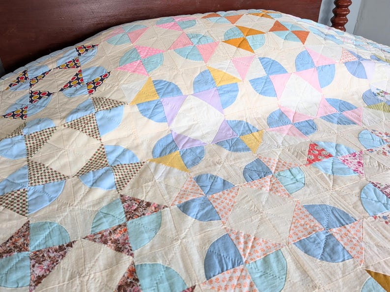 Puede incluir: Un edred&oacute;n hecho a mano con un fondo crema y un patr&oacute;n repetido de formas pastel. El edred&oacute;n presenta arcos y tri&aacute;ngulos azules, rosas, amarillos y naranjas, creando un dise&ntilde;o de patchwork. El edred&oacute;n est&aacute; sobre un marco de cama de madera.