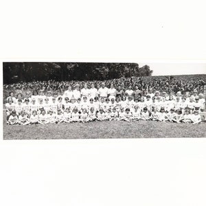May include: Black and white photograph of a large group of children and adults standing in a field. The children are all wearing white shirts and shorts, and the adults are wearing white shirts and pants. The group is standing in front of a field of tall grass.