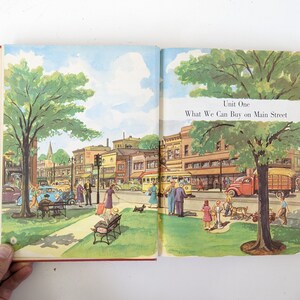 May include: An illustration of a small town Main Street with shops and buildings. There are people walking, cars driving, and a streetcar. The text "Unit One What We Can Buy on Main Street" is at the top of the page.