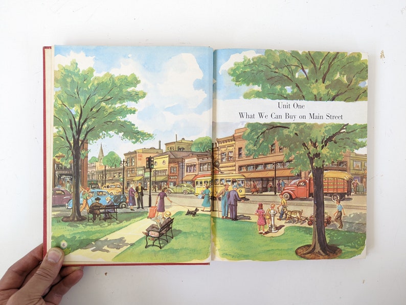 May include: An illustration of a small town Main Street with shops and buildings. There are people walking, cars driving, and a streetcar. The text "Unit One What We Can Buy on Main Street" is at the top of the page.