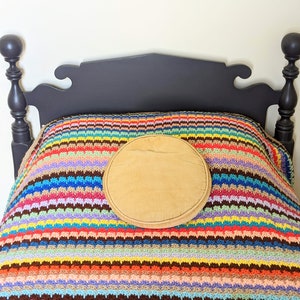Op de afbeelding: Een zwart houten bedframe met een kleurrijk gestreept gehaakt deken en een rond, bruin kussen erop.