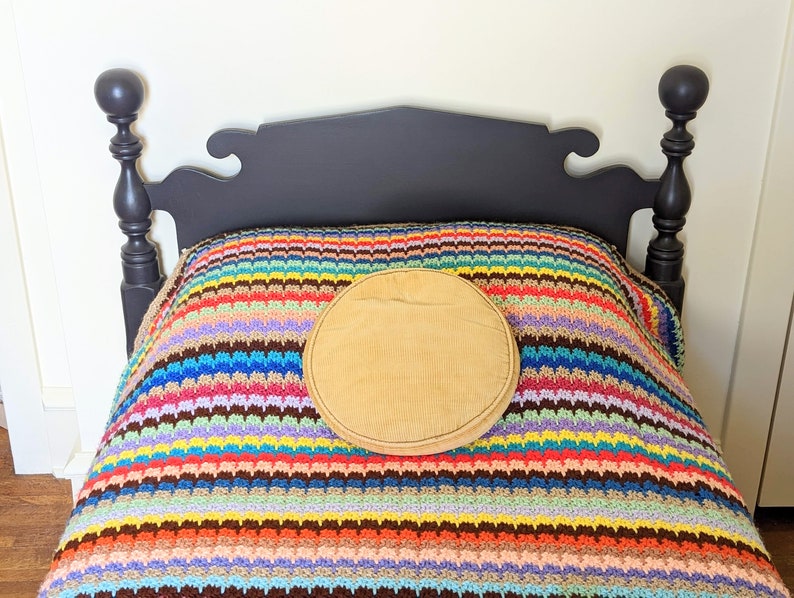 Op de afbeelding: Een zwart houten bedframe met een kleurrijk gestreept gehaakt deken en een rond, bruin kussen erop.