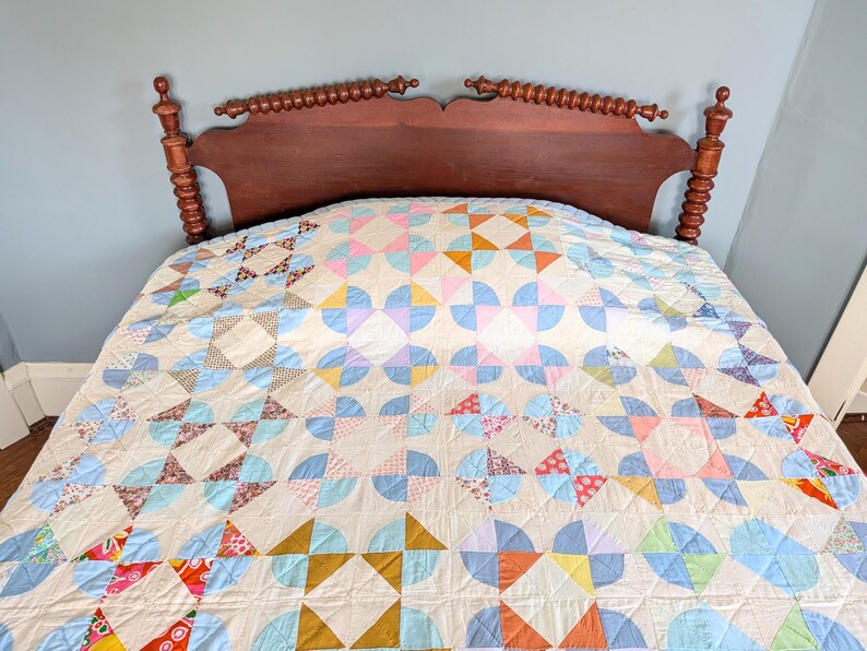 Puede incluir: Un edred&oacute;n hecho a mano con un dise&ntilde;o de patchwork colorido. El edred&oacute;n presenta un patr&oacute;n repetitivo de semic&iacute;rculos y tri&aacute;ngulos en tonos de azul, rosa, naranja y amarillo. El edred&oacute;n est&aacute; colocado sobre una cama con un cabecero de madera oscura.