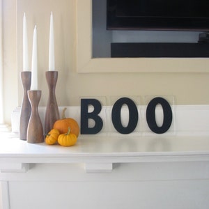 May include: A white fireplace mantel with a black BOO sign, three wooden candle holders with white candles, and three small orange pumpkins.