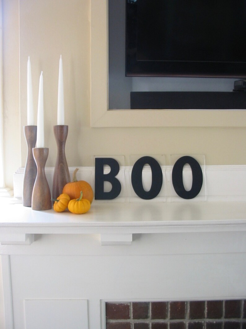 May include: A white fireplace mantel with a black BOO sign, three wooden candle holders with white candles, and three small orange pumpkins.