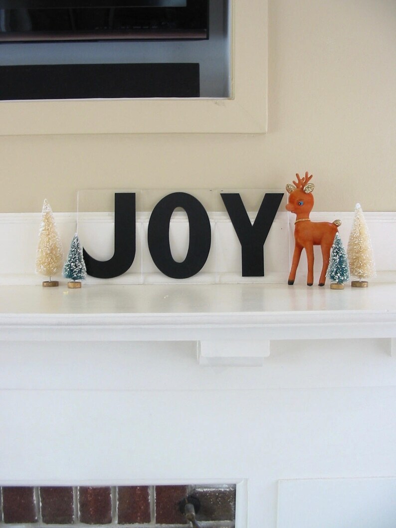May include: Black wooden letters spelling "JOY" on a white mantel with two small artificial pine trees and a red reindeer figurine.