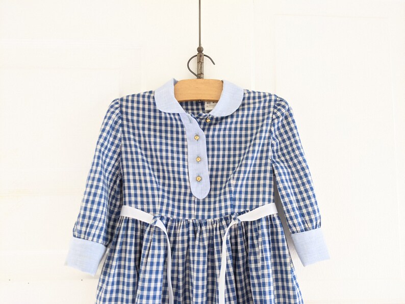 Op de afbeelding: Een blauw-wit geruit jurkje met een witte kraag en een witte strik. Het jurkje heeft lange mouwen met blauw-wit geruite manchetten.
