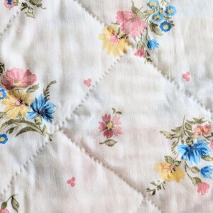 Puede incluir: Primer plano de una tela acolchada blanca con un estampado floral. El estampado presenta flores rosas, amarillas y azules con hojas verdes. El acolchado crea un patr&oacute;n de diamantes en la tela.