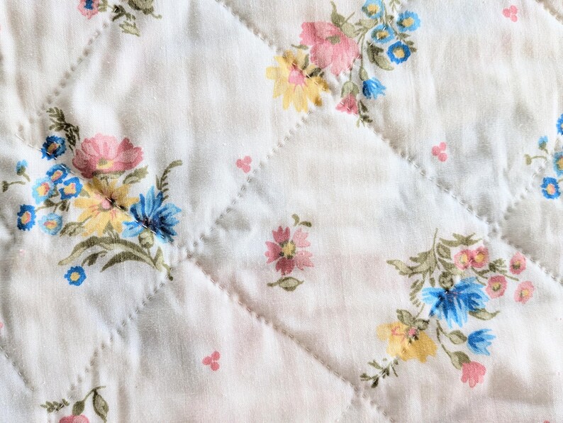 Puede incluir: Primer plano de una tela acolchada blanca con un estampado floral. El estampado presenta flores rosas, amarillas y azules con hojas verdes. El acolchado crea un patr&oacute;n de diamantes en la tela.