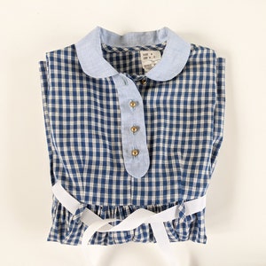 Op de afbeelding: Een blauw-wit geruit jurkje met een witte kraag en een witte strik. Het jurkje heeft een knoopsluiting en een label met de tekst "SIZE 4, LEFT P, STYLE 1".